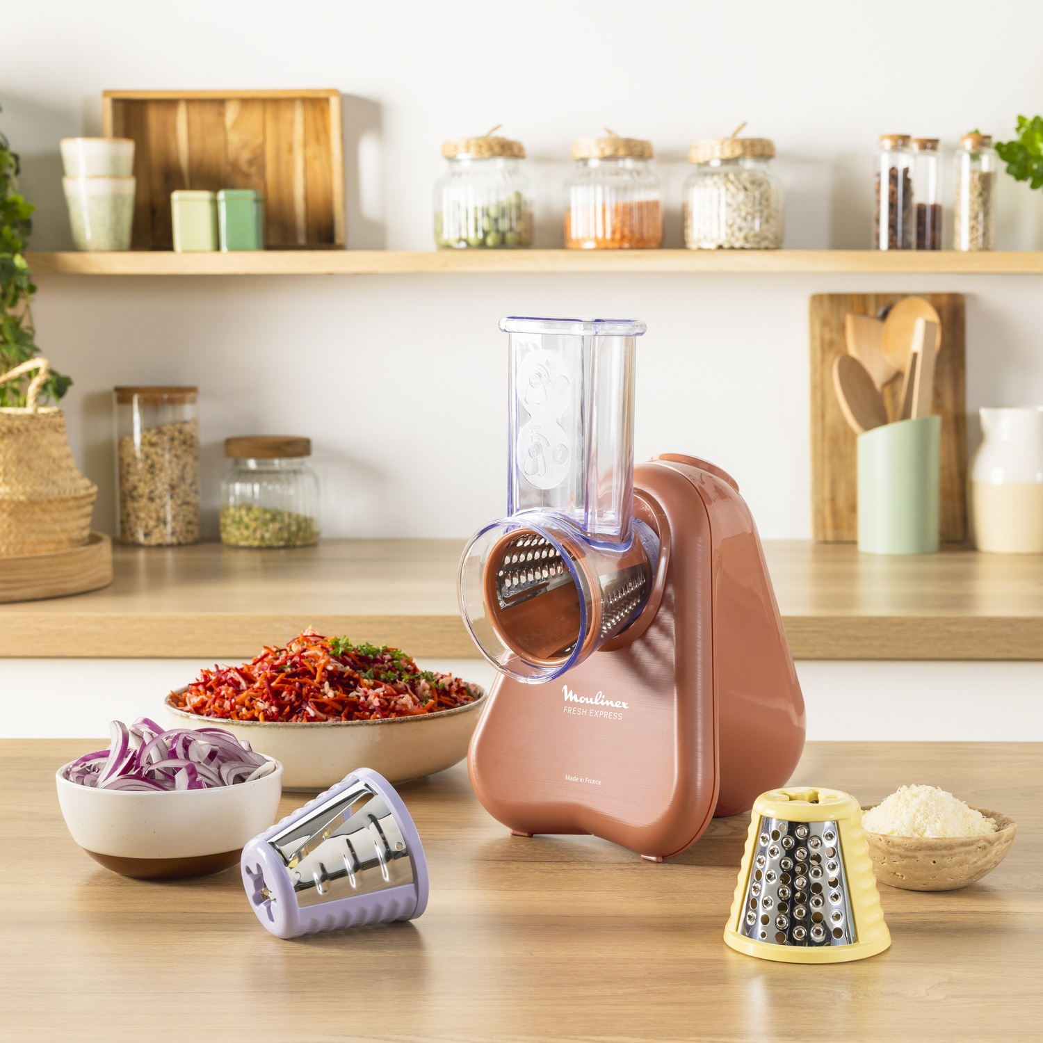 A Moulinex Fresh Express, bowls of vegetables, and a bowl of red onion on a wooden counter. Jars and shelves are in the background.