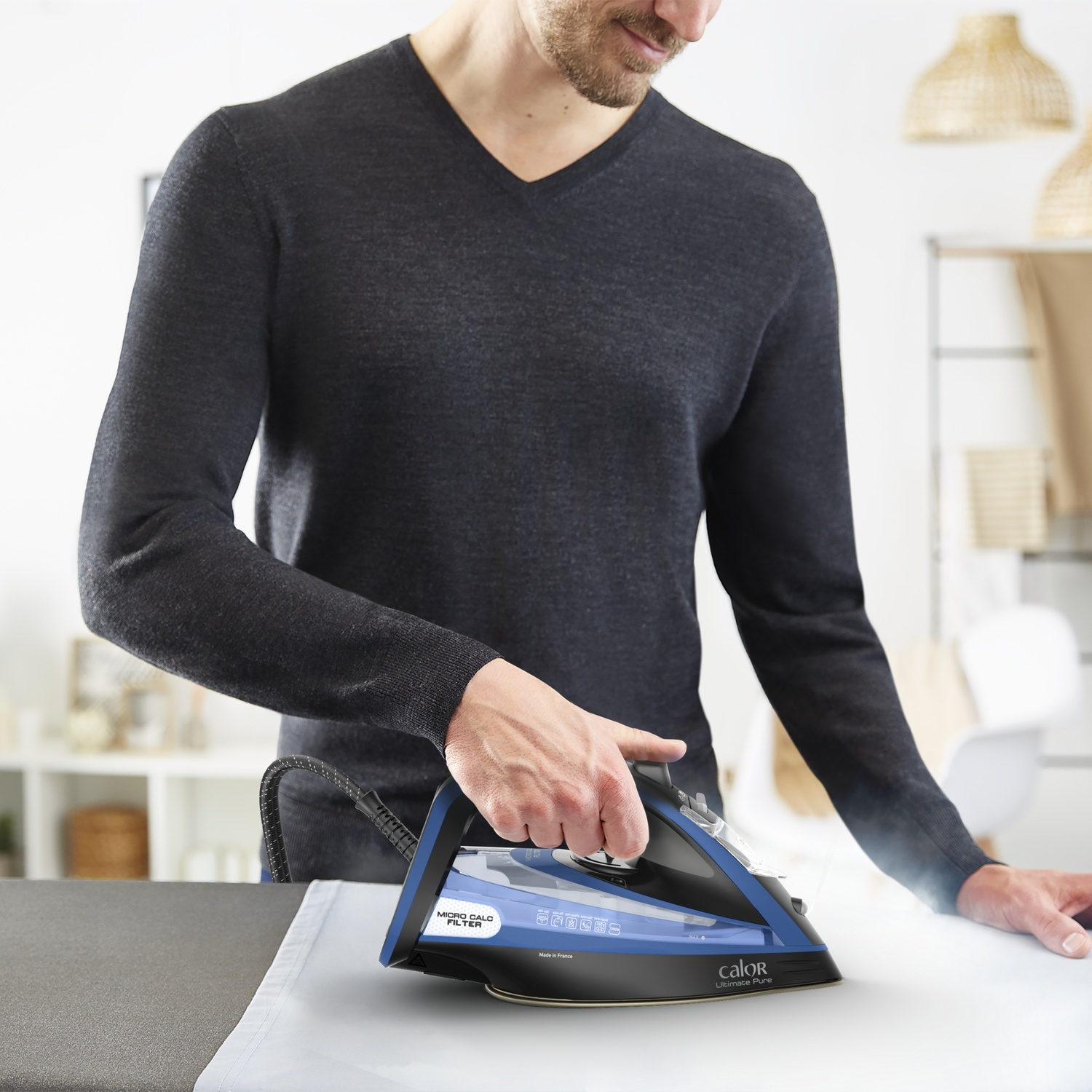Un homme repasse une chemise blanche avec un fer à repasser bleu et noir. De la vapeur s'élève. Il porte un pull sombre.