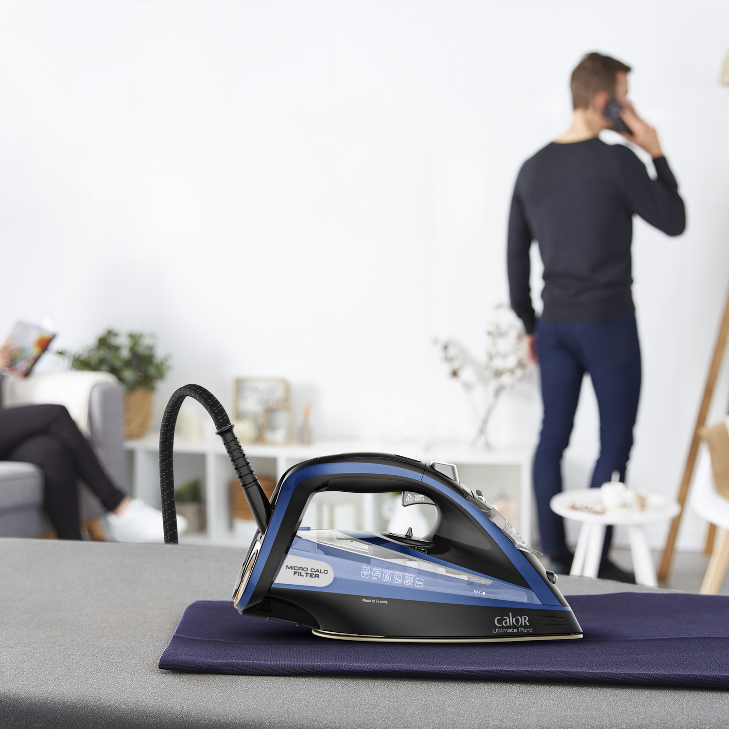 Un fer à repasser avec des touches de bleu sur un tissu foncé, une personne au téléphone en arrière-plan.