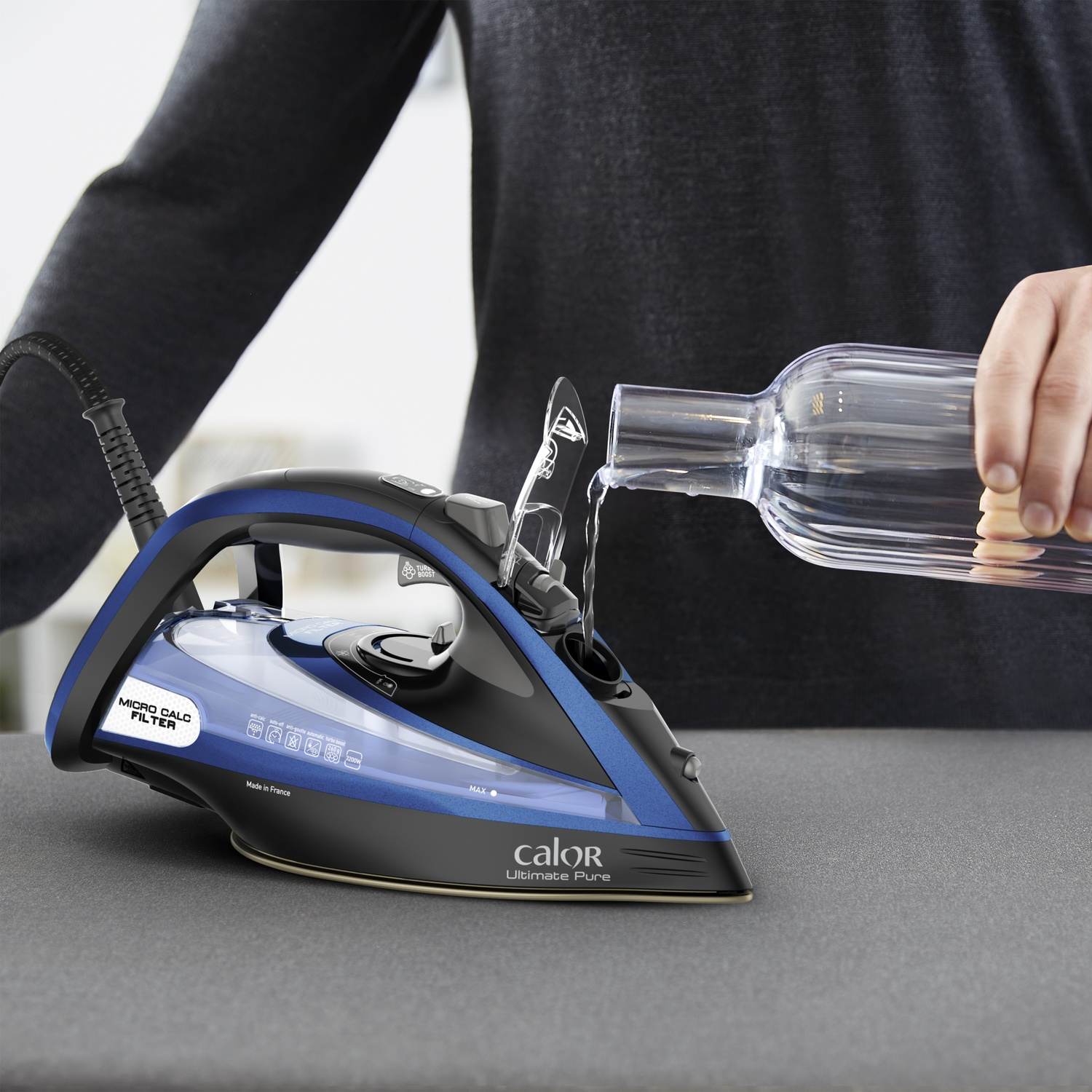 Une personne verse de l'eau dans un fer à repasser bleu et noir. Le fer est sur une surface grise, avec une bouteille transparente.