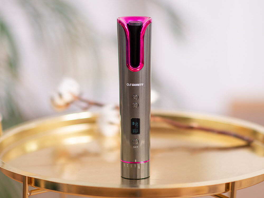 Lokówka z różowym akcentem stoi na złotym stoliku. Jest szara, z cyfrowym wyświetlaczem. Tło z rozmytymi kwiatami.