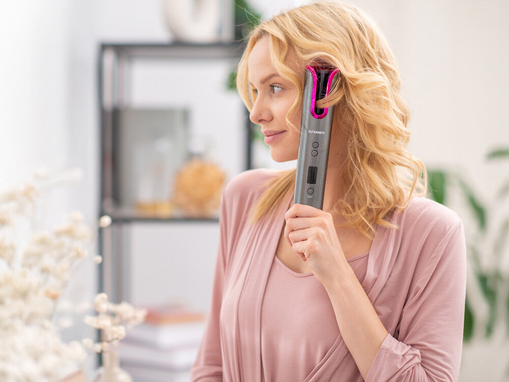 Blondynka używa lokówki. Ma na sobie różowy top i szlafrok. Na tle półka i kwiaty.