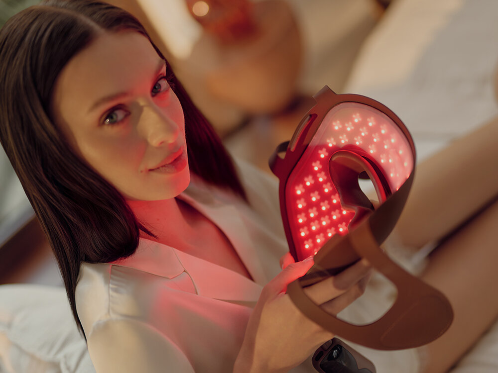 Kobieta trzymająca maskę do terapii światłem czerwonym. Ma na sobie białą koszulę i długie, ciemne włosy. Tło jest rozmyte.