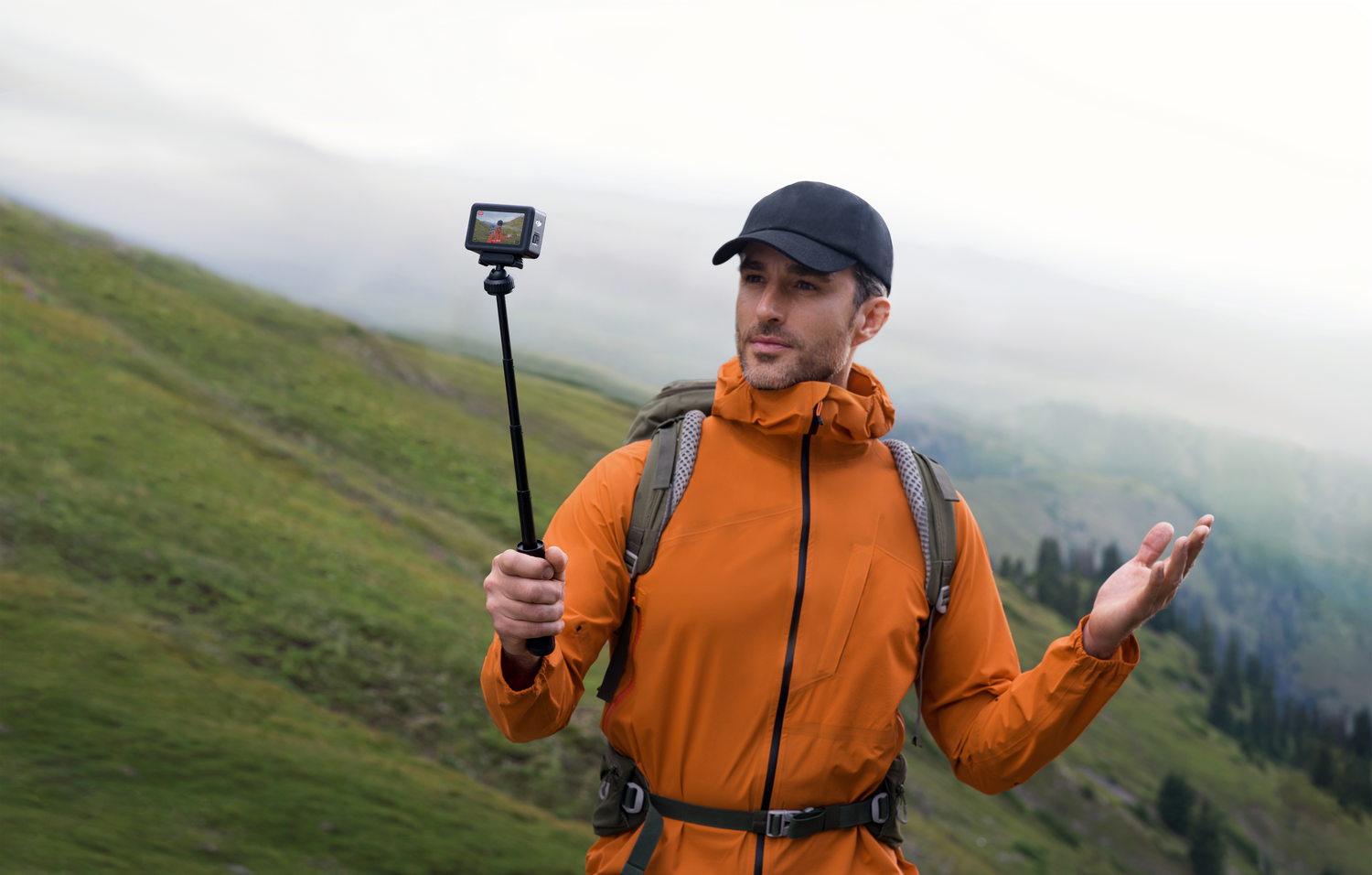 Hombre con chaqueta naranja sosteniendo cámara en un palo selfie en montaña. Gesticula con las manos.