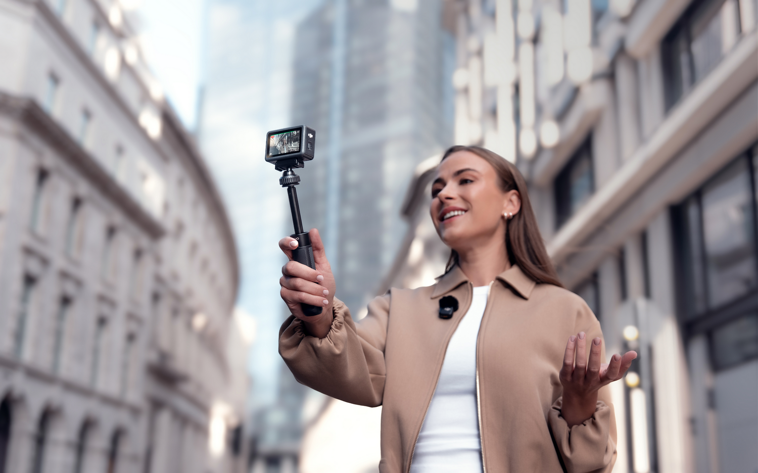 Una mujer sostiene una cámara en un palo de selfie, sonriendo en un entorno urbano. Edificios al fondo. Lleva una chaqueta color canela.