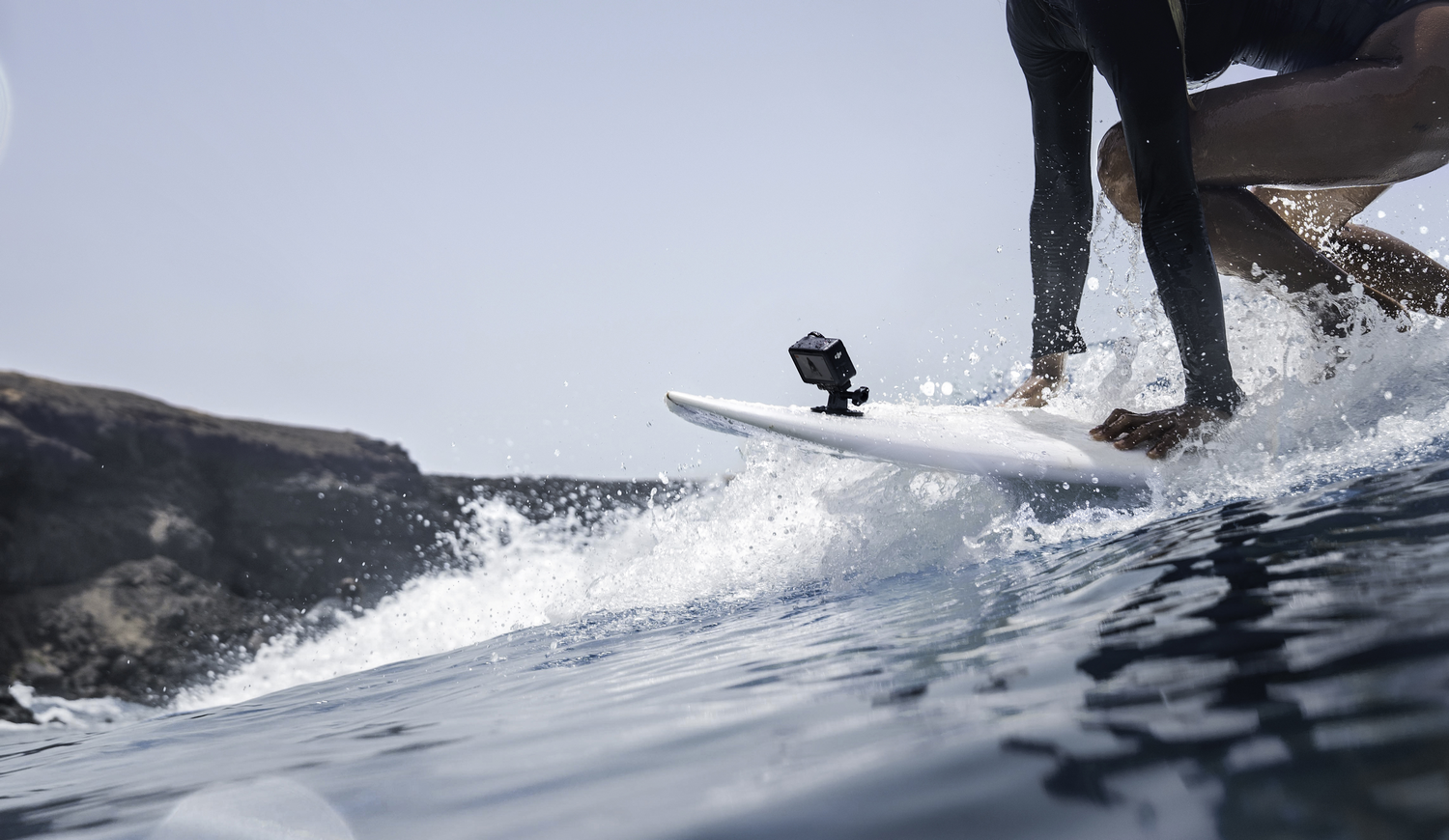 Un surfista surfea una ola. El surfista está en una tabla de surf blanca con una cámara. El agua salpica.