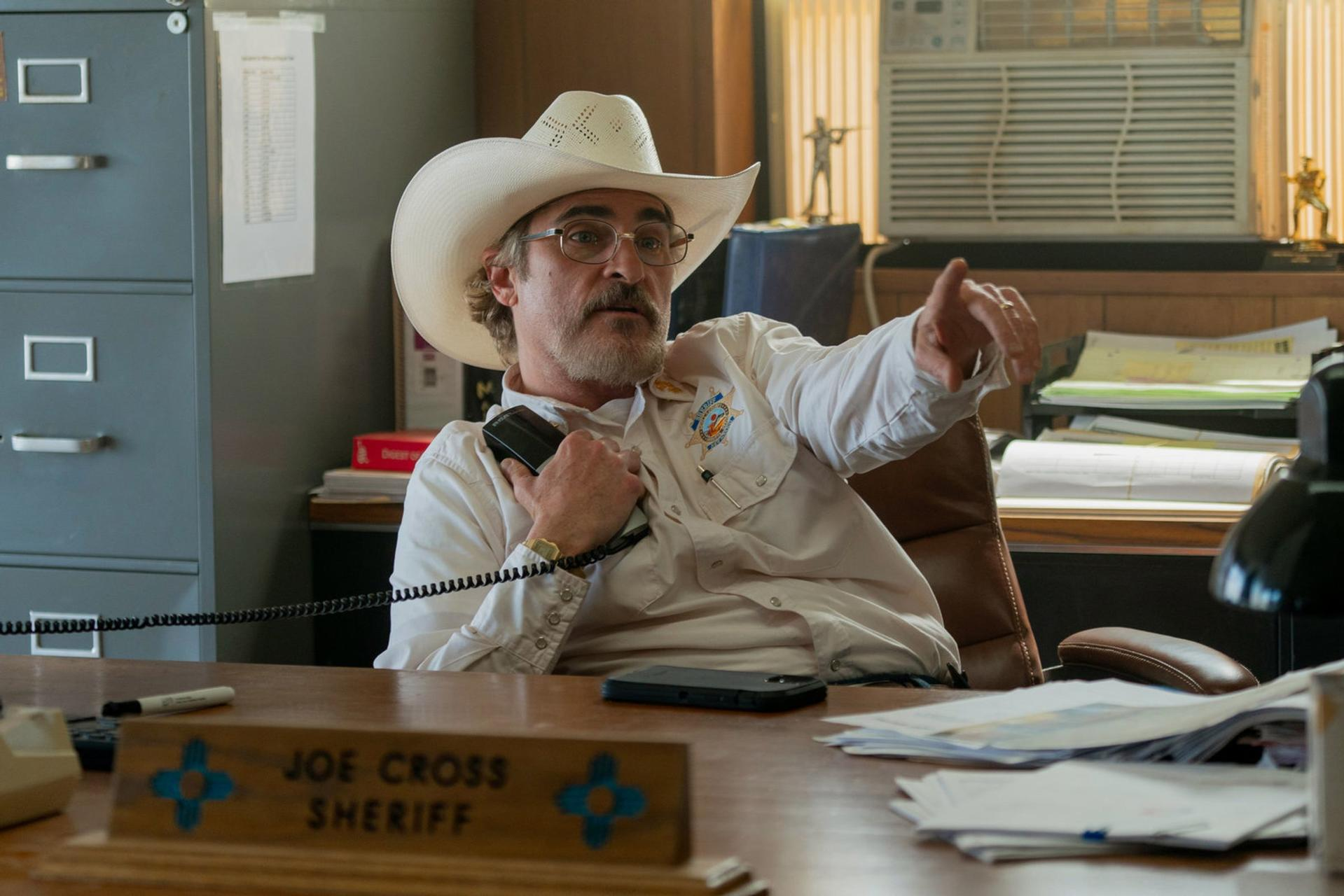 Sheriff mit Cowboyhut telefoniert und zeigt. Büro mit Aktenschrank, Schreibtisch mit Papieren und Auszeichnungen.