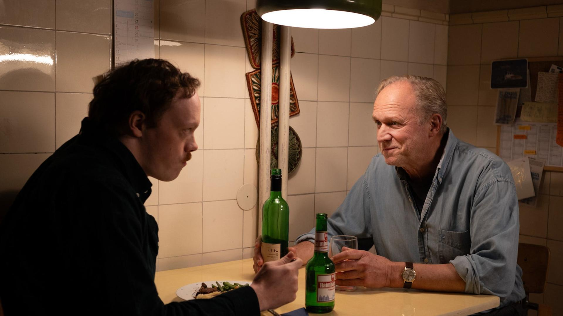 Zwei Männer sitzen an einem Tisch, Flaschen und Gläser vorhanden. Einer ist jünger, der andere älter. Sie sind in einem kleinen Raum.