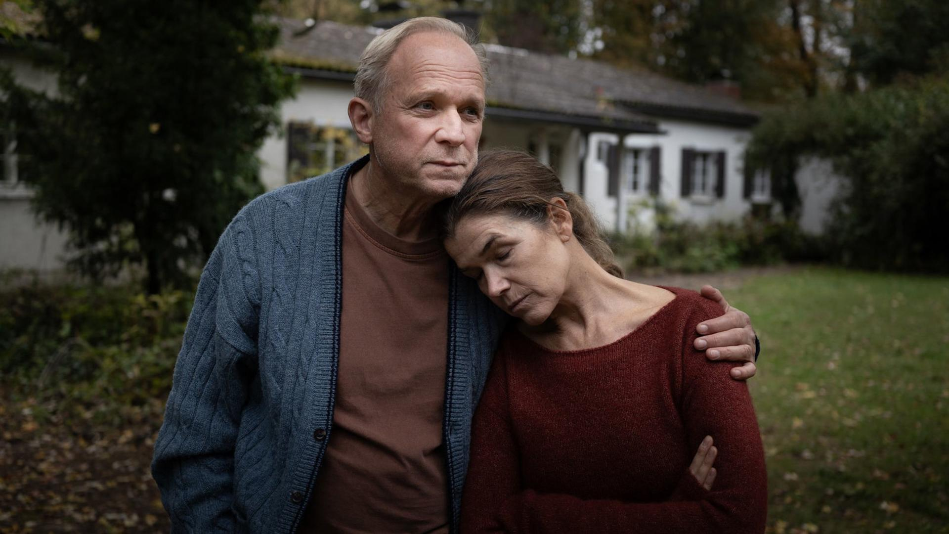 Ein älterer Mann in blauem Cardigan umarmt eine Frau in rotem Pullover. Sie stehen vor einem Haus. Die Szene hat eine melancholische Stimmung.