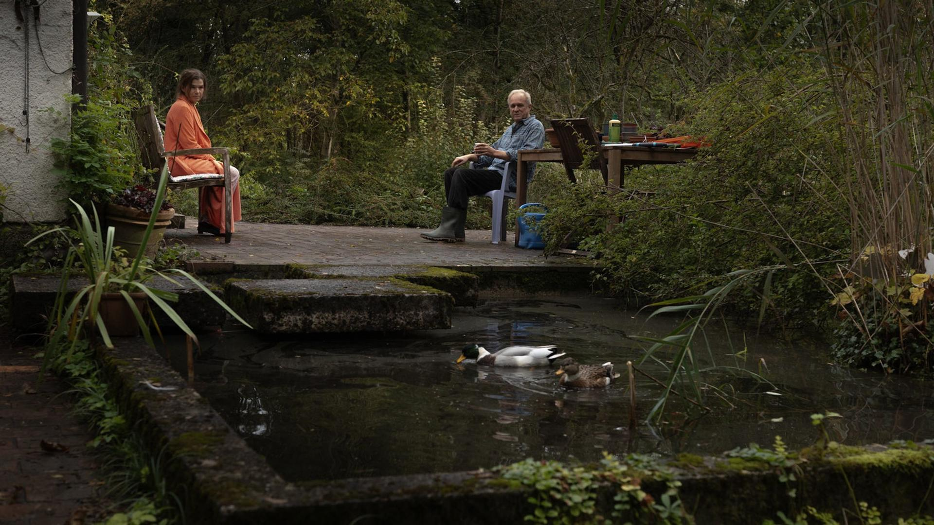 Zwei Personen sitzen draußen in der Nähe eines Teichs mit Enten. Bäume umgeben die Gegend. Im Hintergrund stehen ein Tisch und Stühle.