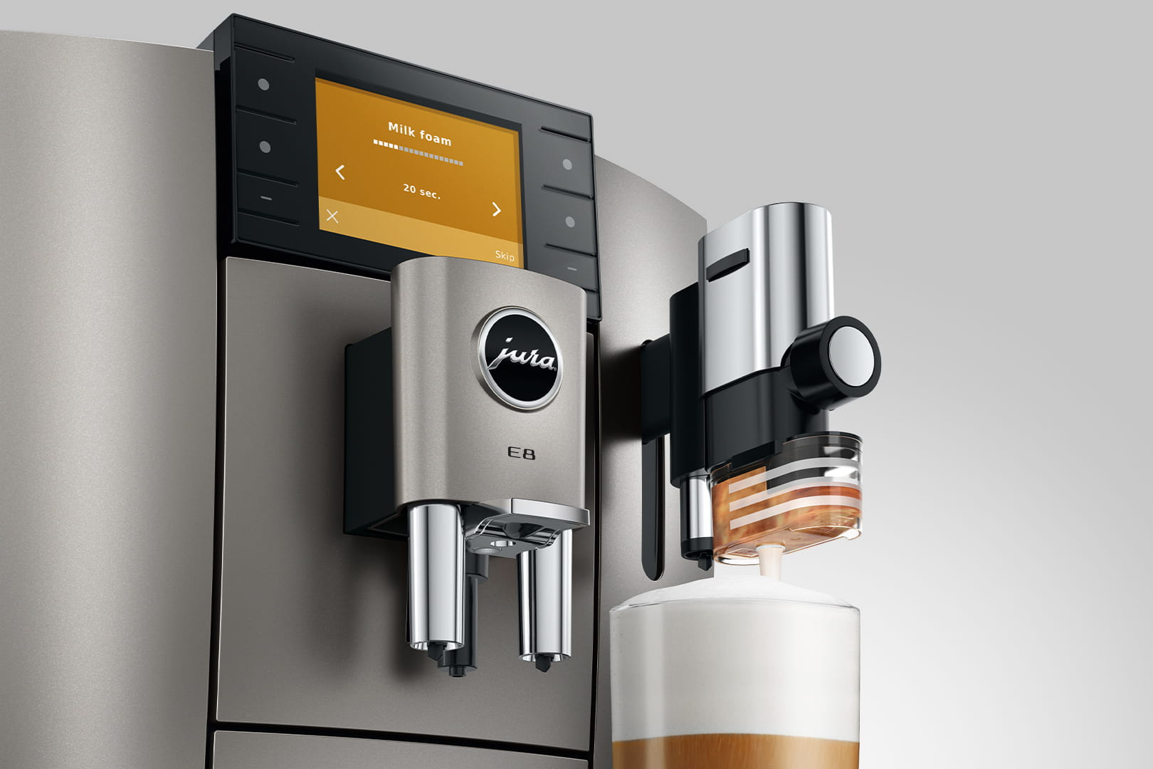 Una macchina da caffè Jura E8 che produce schiuma di latte. Lo schermo mostra 'Schiuma di latte' e un timer. Un bicchiere si riempie di caffè e schiuma.