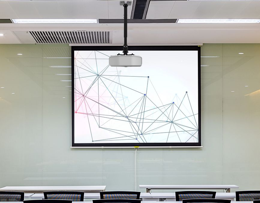 Ein Projektor hängt von der Decke und projiziert eine abstrakte Grafik auf eine Leinwand. Tische und Stühle befinden sich im Vordergrund, in einem Klassenzimmer.