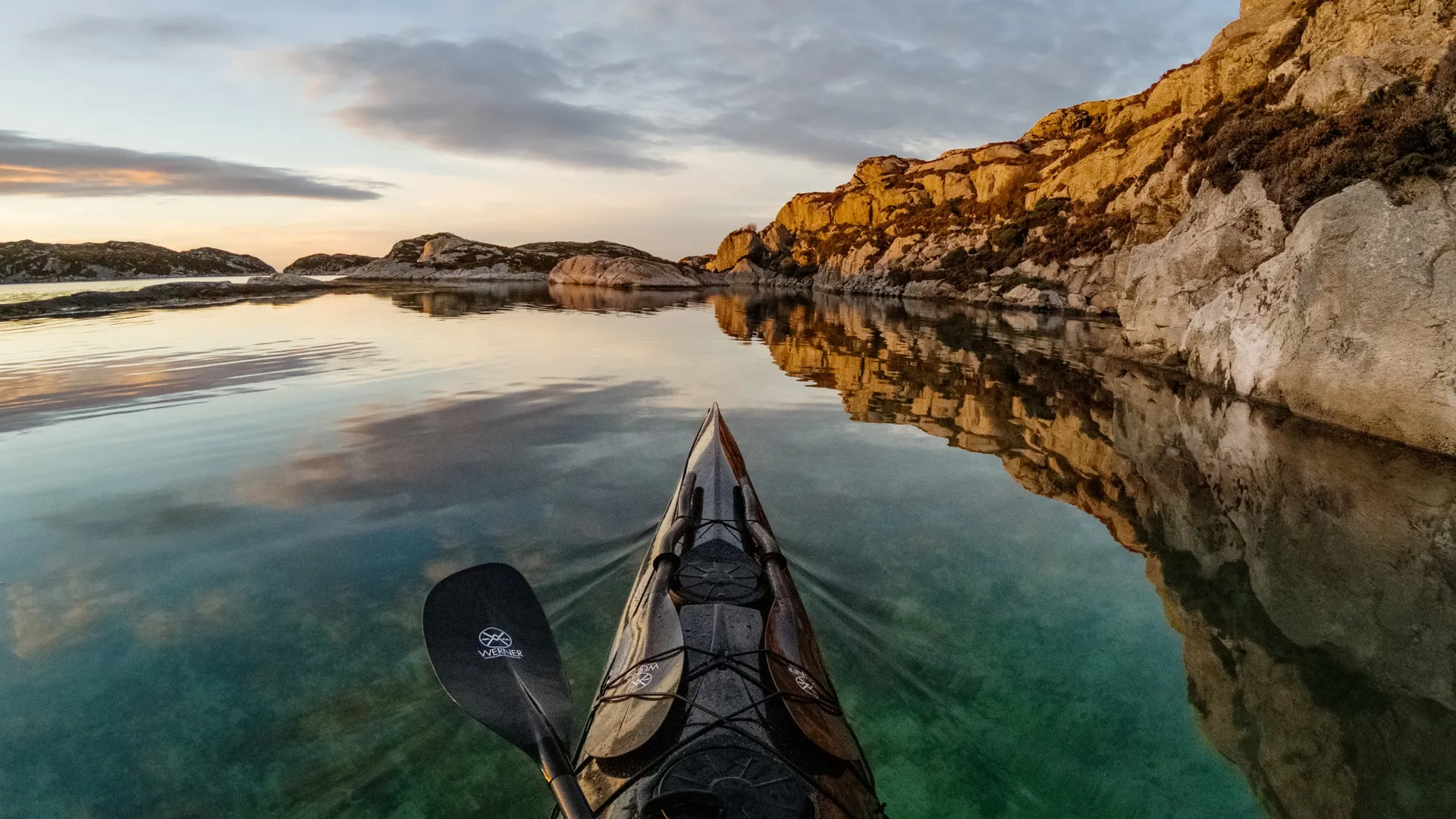 Kajak auf ruhigem Wasser, Paddel im Blick. Felsenküsten und Wolken spiegeln sich im Wasser. Goldenes Licht rechts.