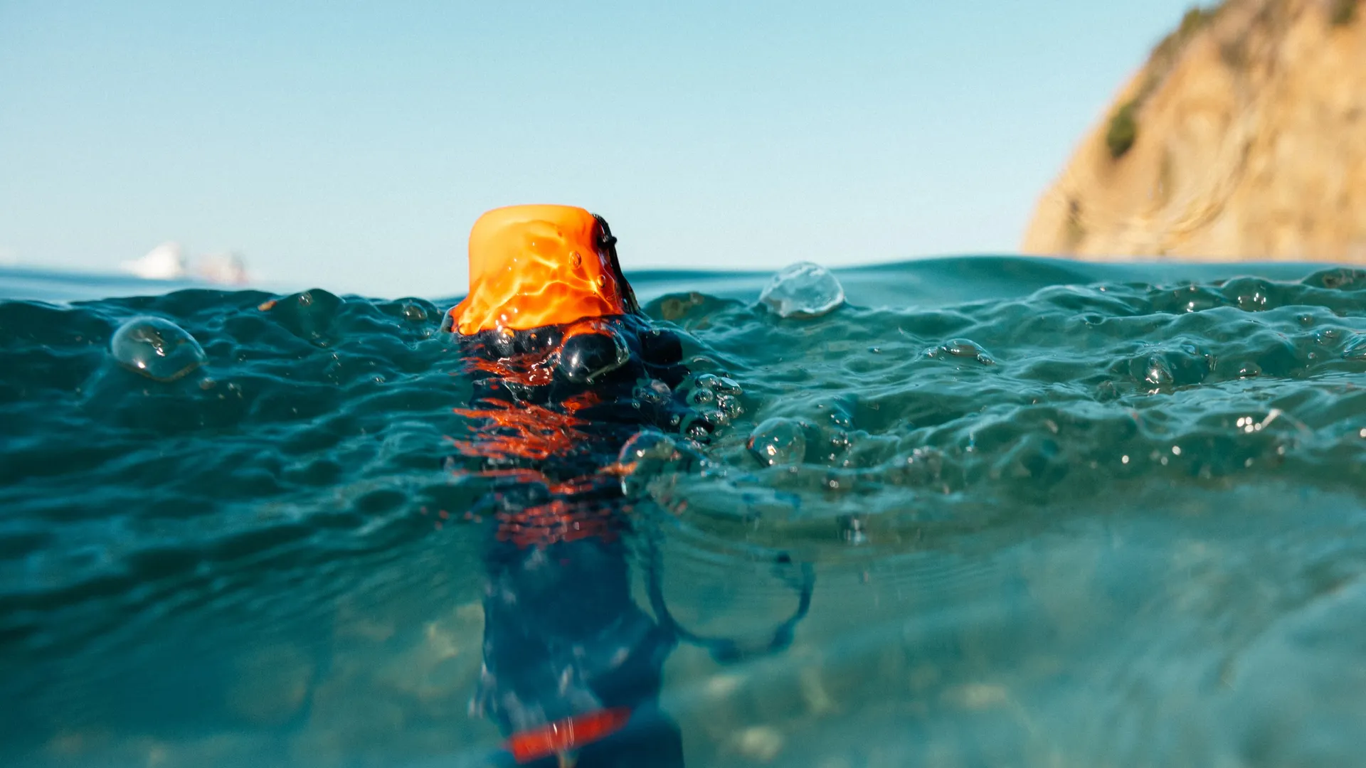 Ein orangefarbenes Objekt schwimmt im Wasser. Blasen umgeben es. Das Meer ist blaugrün, mit klarem Himmel und einer Klippe im Hintergrund.