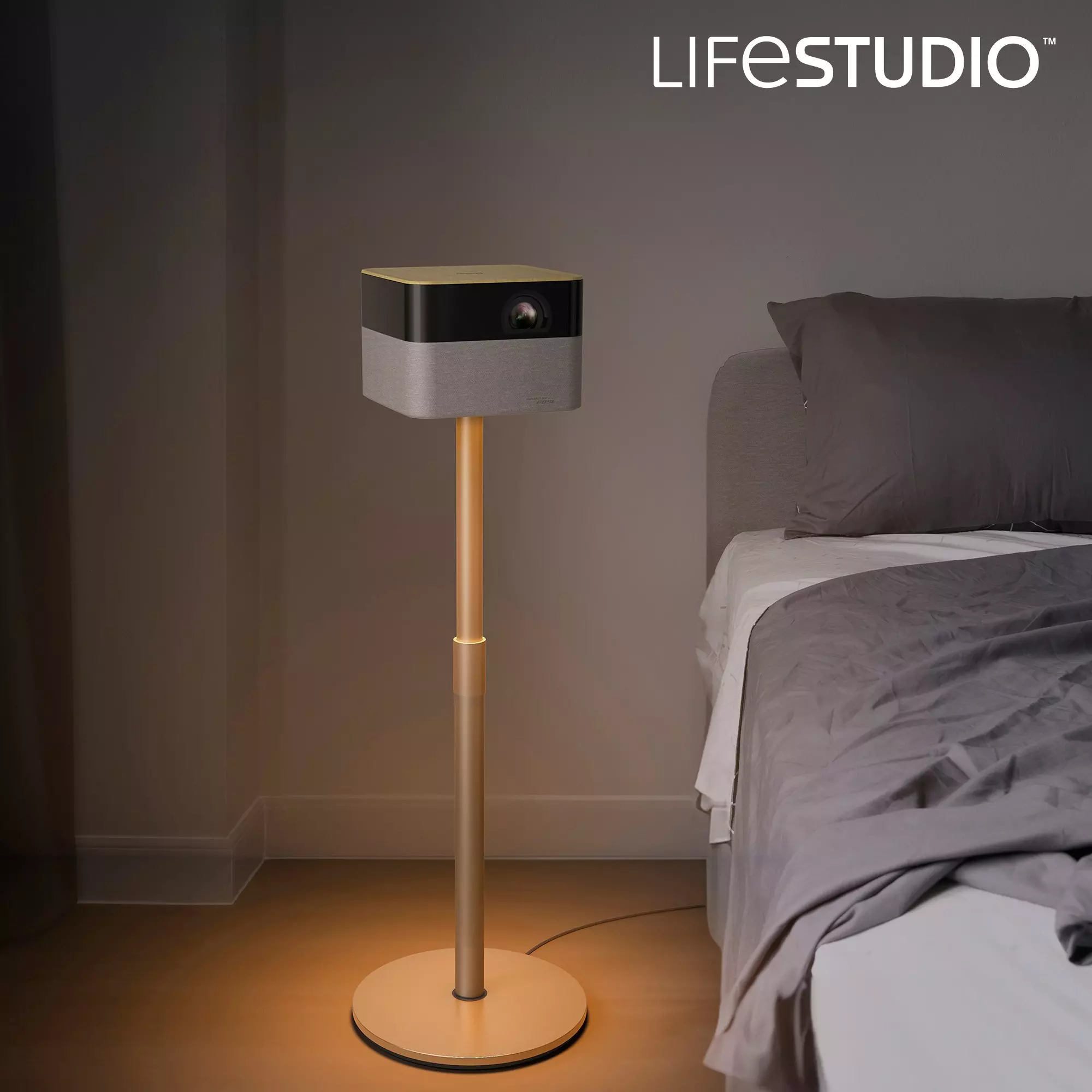 Un proiettore su un supporto accanto a un letto. Il proiettore ha la parte superiore dorata e il corpo grigio. Il letto ha la biancheria da letto grigia.