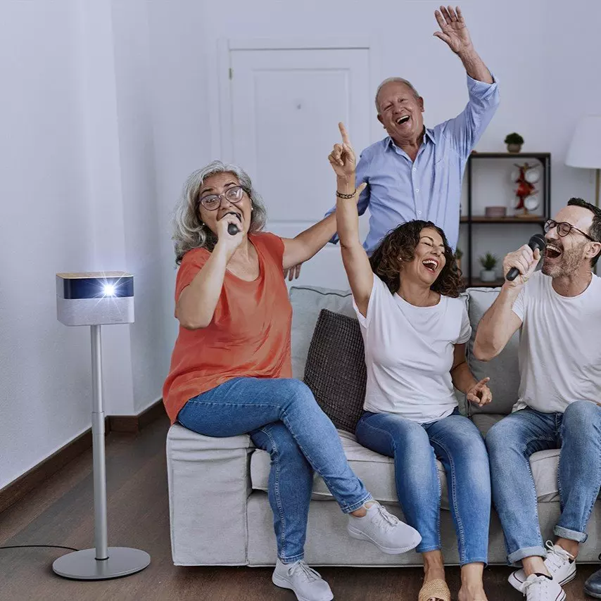 Quattro persone cantano con microfoni, ridendo. Un proiettore a sinistra. Sono seduti su un divano grigio in soggiorno.