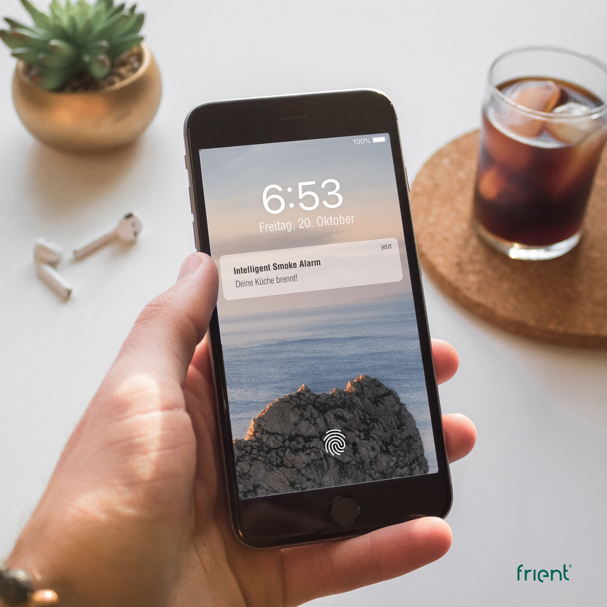 Hand, die ein Telefon mit einer Rauchmelder-Benachrichtigung hält. Eine Pflanze, Ohrhörer und ein Getränk befinden sich in der Nähe.