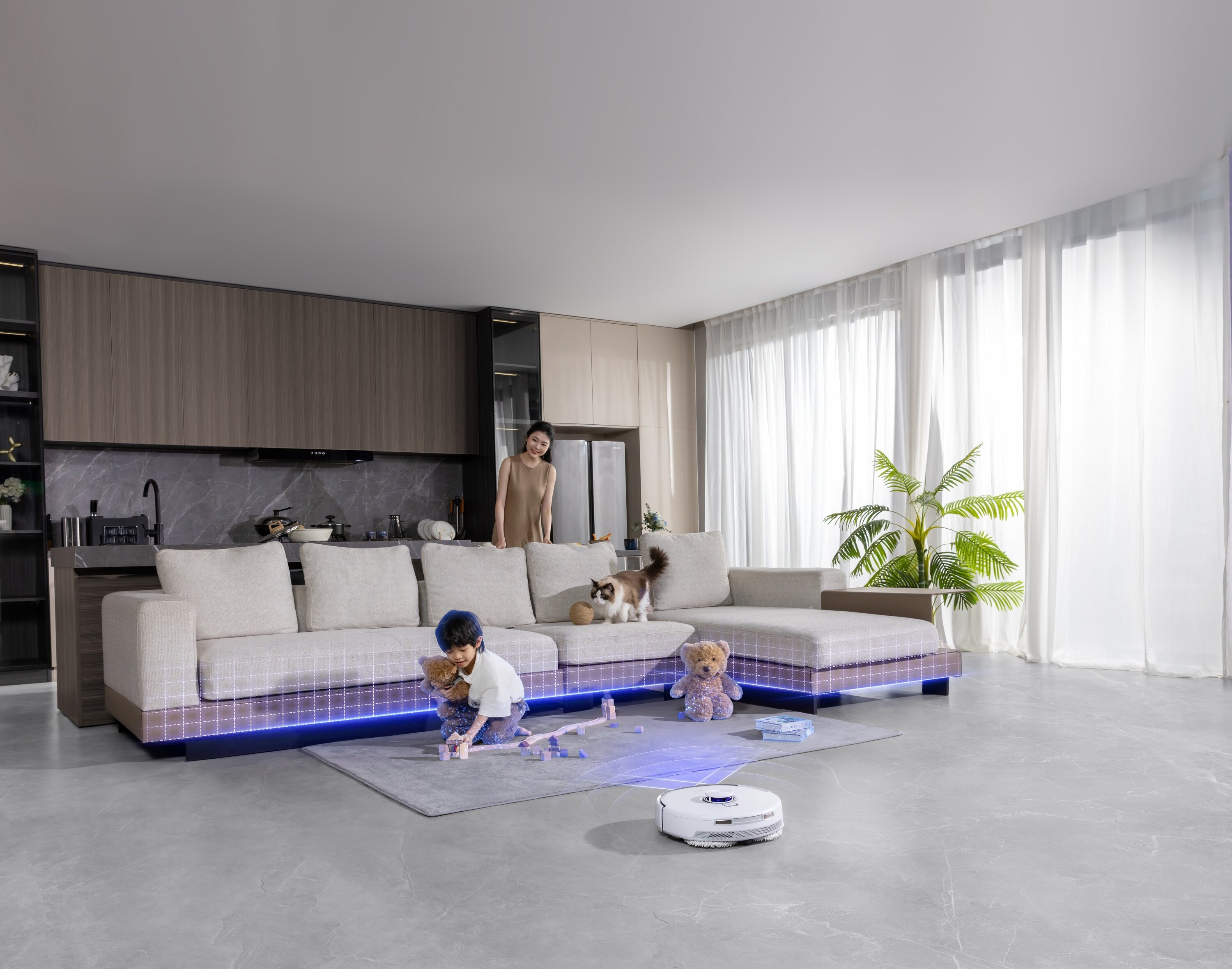 Scena w salonie. Dziecko bawi się zabawkami w pobliżu sofy, robot sprząta podłogę. Obecna jest również kobieta i kot.