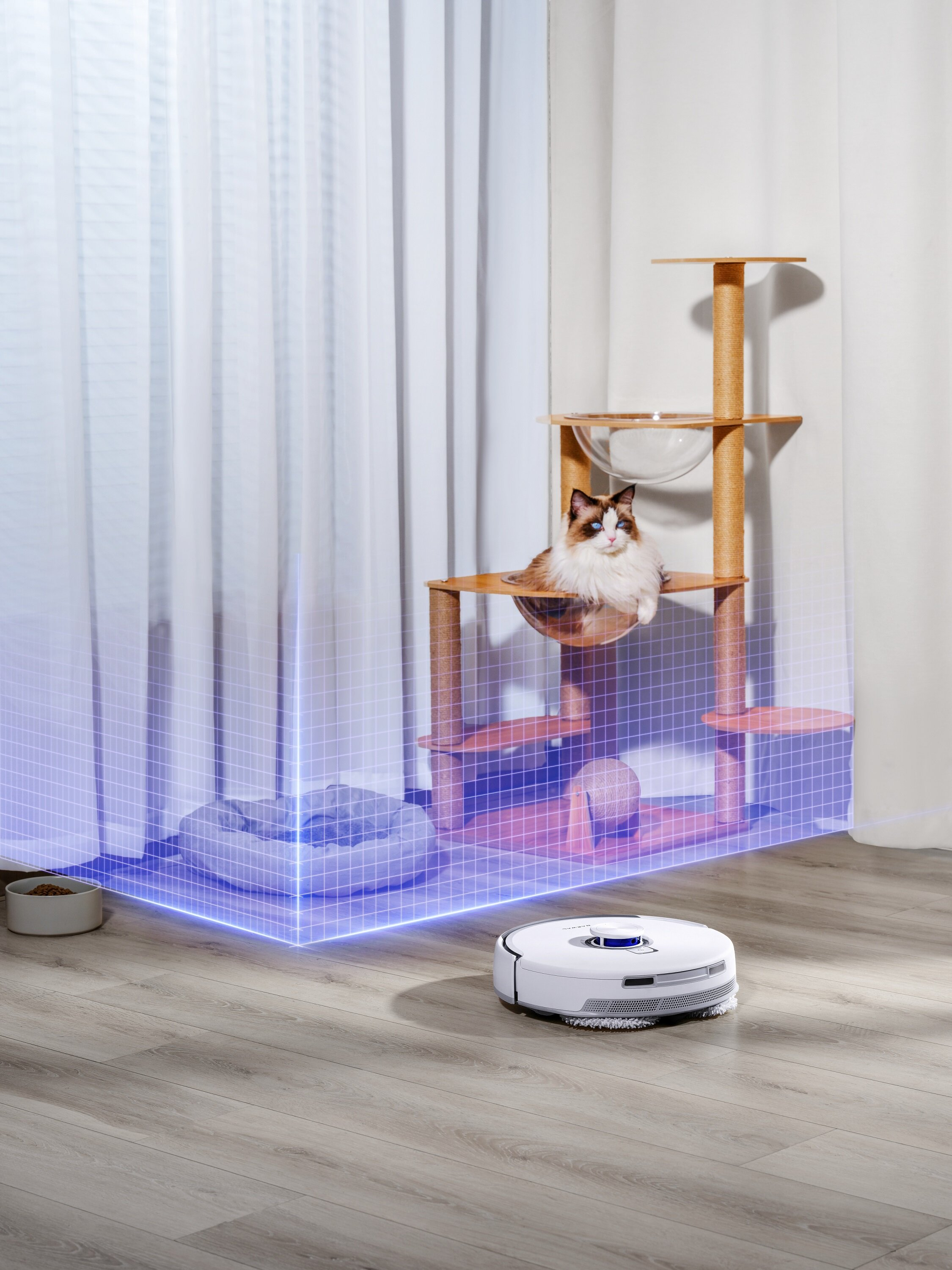 Kot siedzi na drzewie dla kota. Robot sprzątający czyści drewnianą podłogę. Widoczne legowisko dla kota.