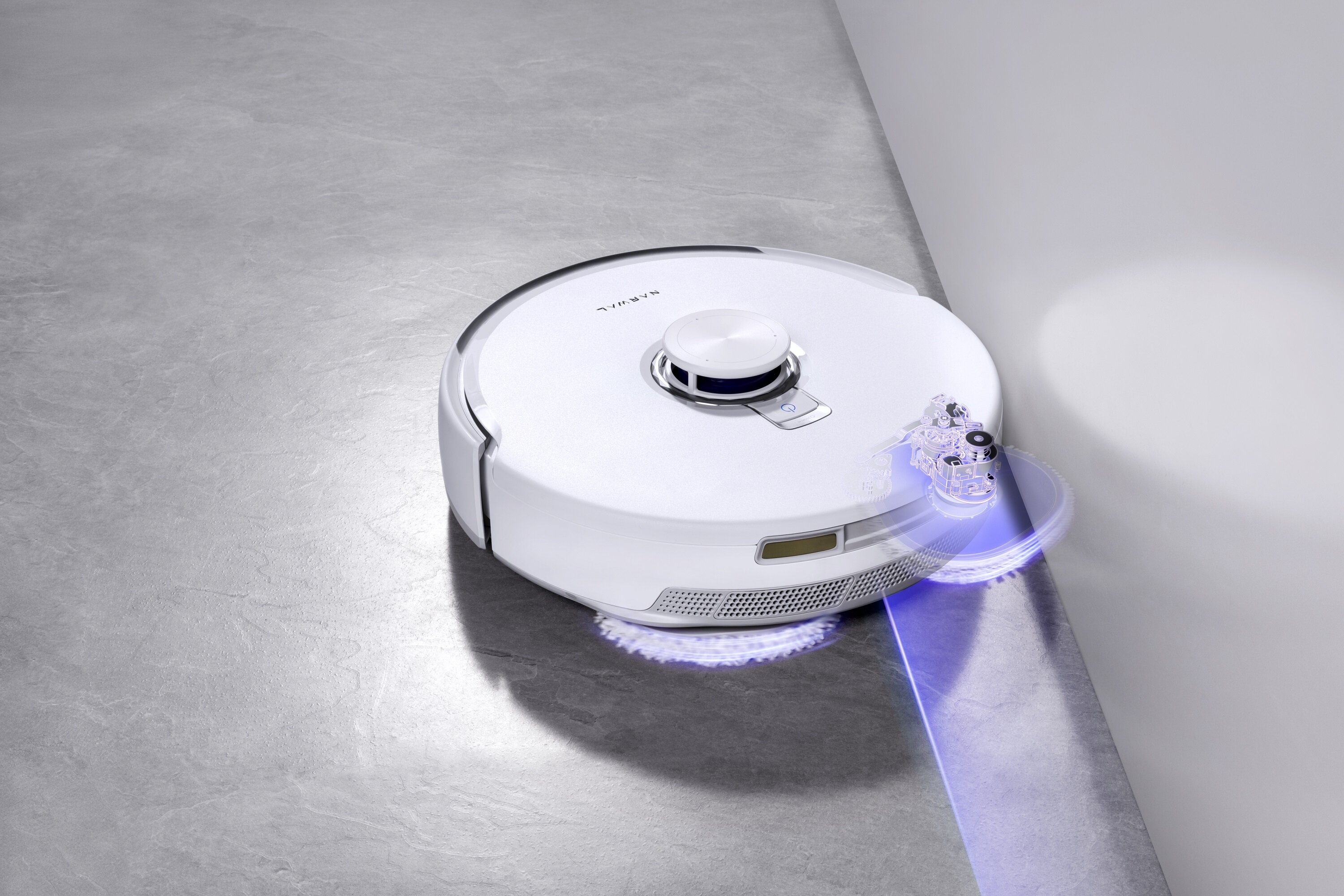 Biały robot odkurzający czyści szarą podłogę obok białej ściany. Robot jest okrągły.