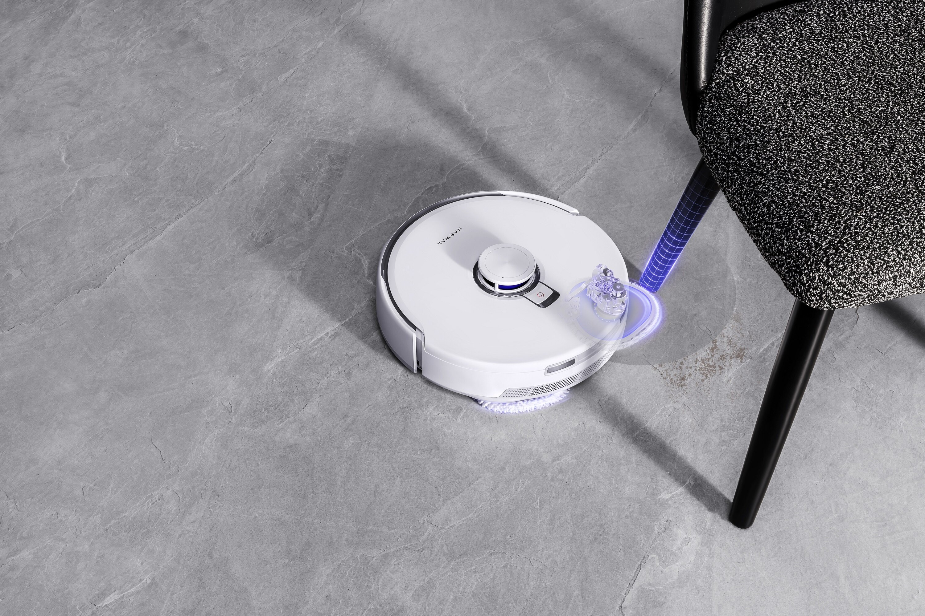 Biały robot odkurzający znajduje się w pobliżu czarnego krzesła. Podłoga jest szara. Niebieskie światło otacza odkurzacz.