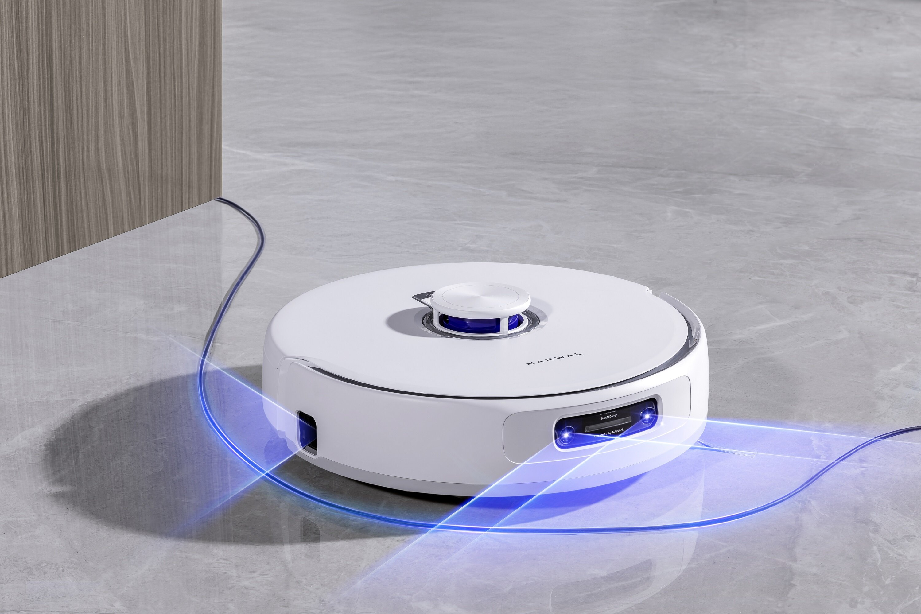 Biały robot odkurzający na szarej podłodze z płytek. Niebieskie światło wokół niego. Drewniana ściana z boku.