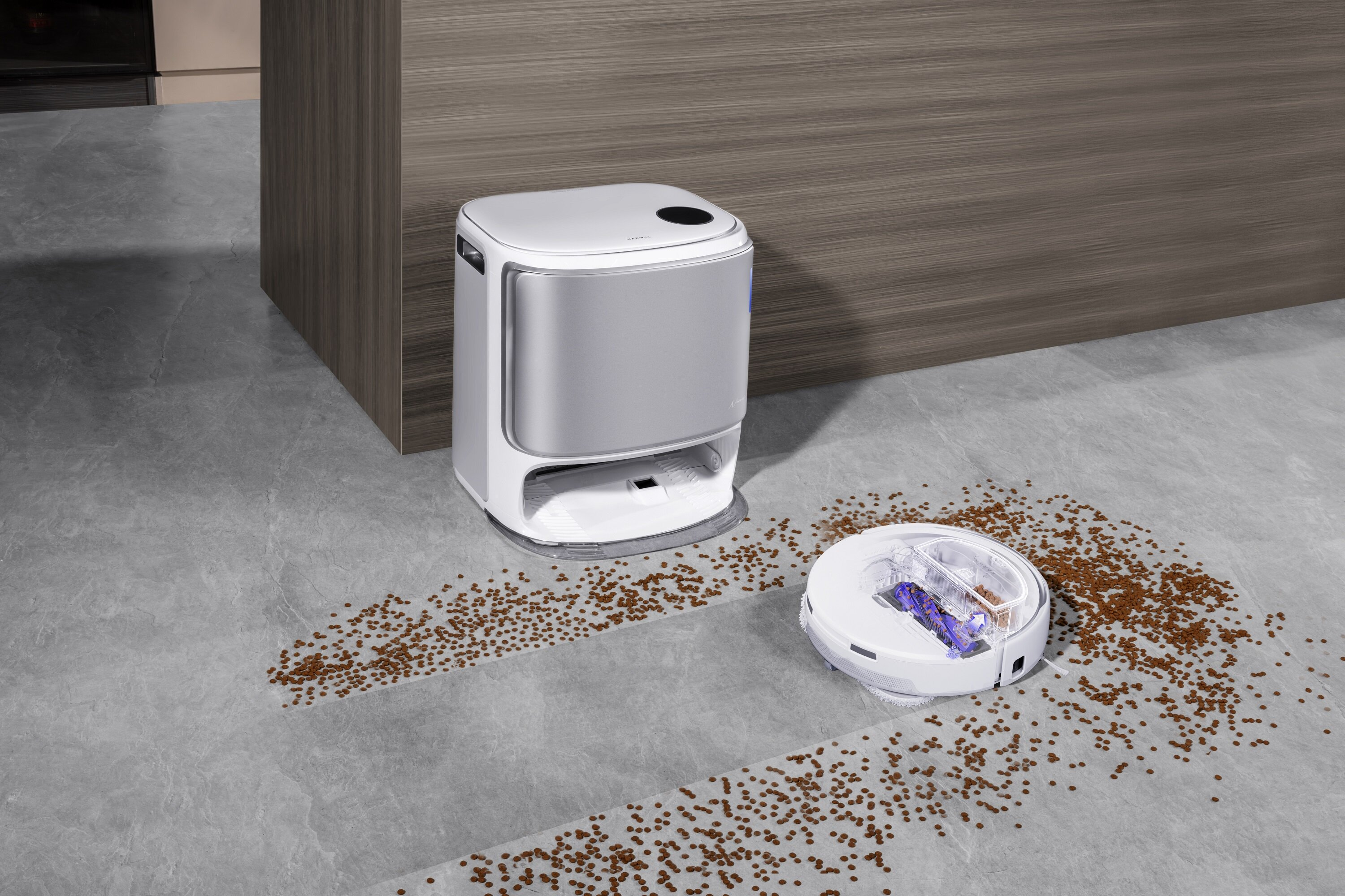 Biały robot odkurzający i stacja ładująca na szarej podłodze. Brązowe śmieci są rozrzucone w pobliżu.