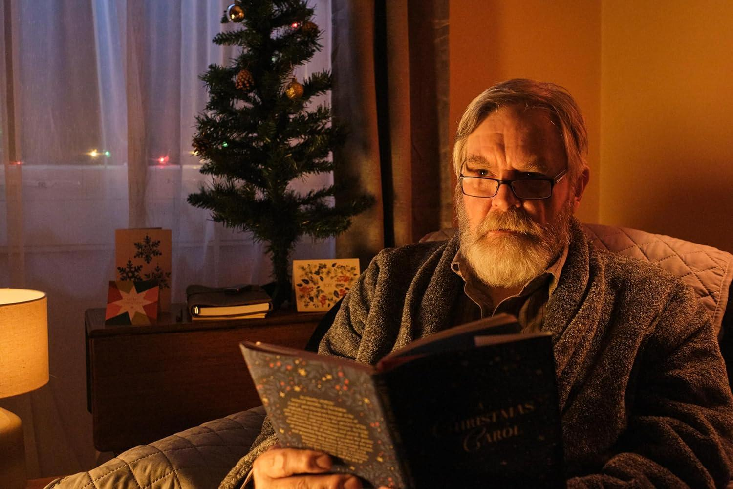 Ein älterer Mann mit Brille liest ein Buch, 'Christmas Carol', neben einem geschmückten Baum und Karten. Er trägt einen Bademantel.