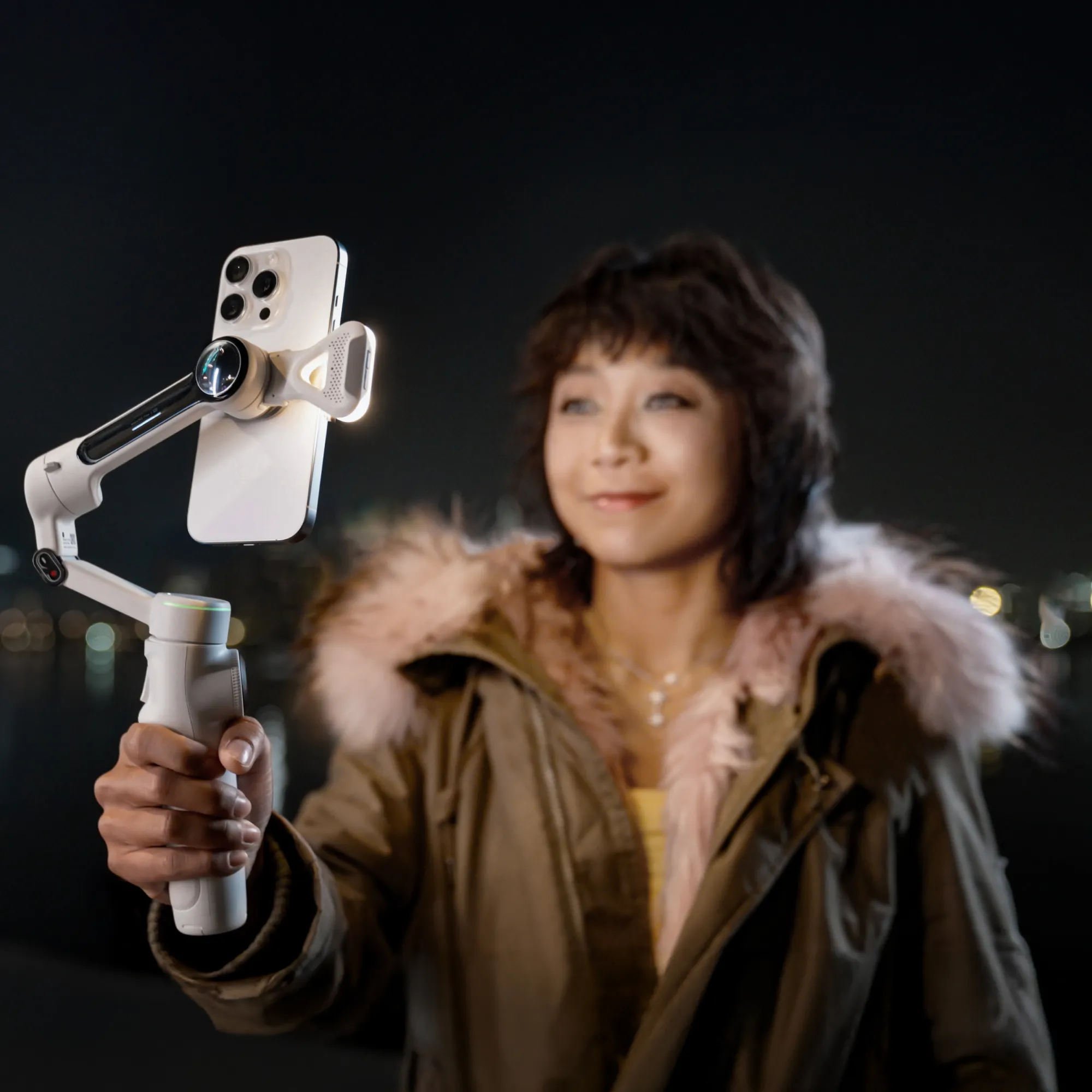 Frau hält ein Telefon auf einem Gimbal und nimmt nachts ein Video auf. Sie trägt eine Jacke mit rosa Pelzkragen.