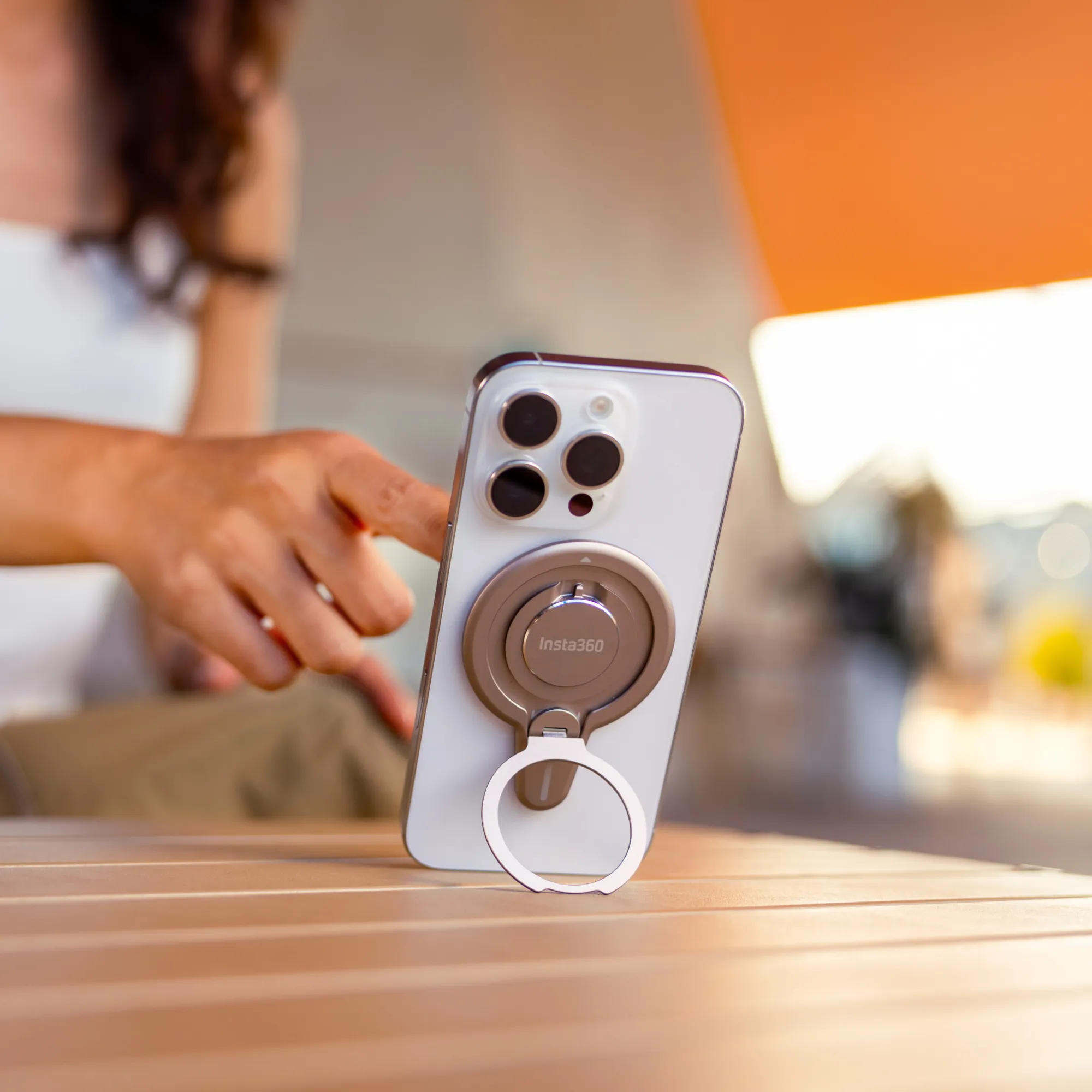 Eine Hand berührt ein Telefon mit einem Insta360-Ringhalter auf einem Holztisch. Der Hintergrund ist unscharf.
