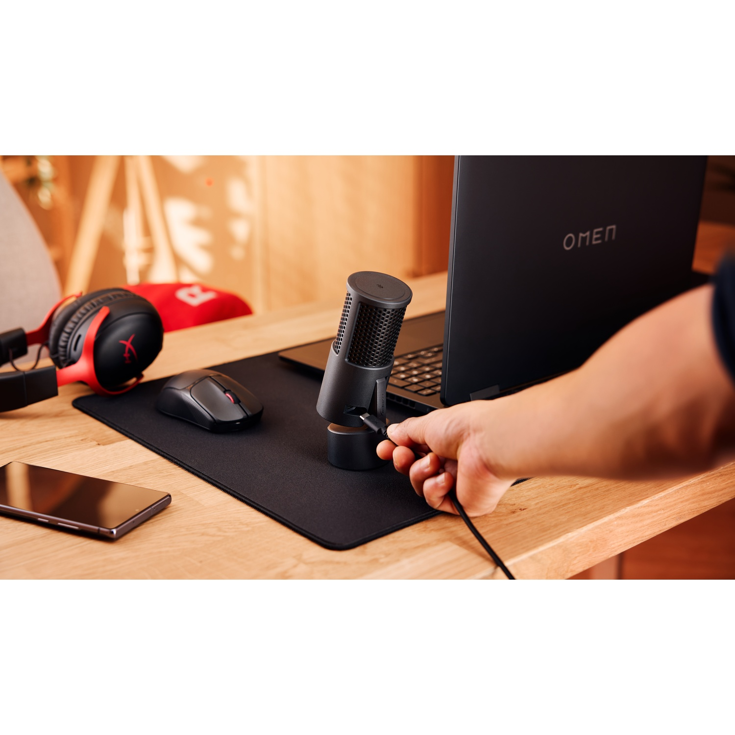 Une personne branche un microphone sur un ordinateur portable noir. Une souris noire, un casque et un téléphone sont sur un bureau en bois.