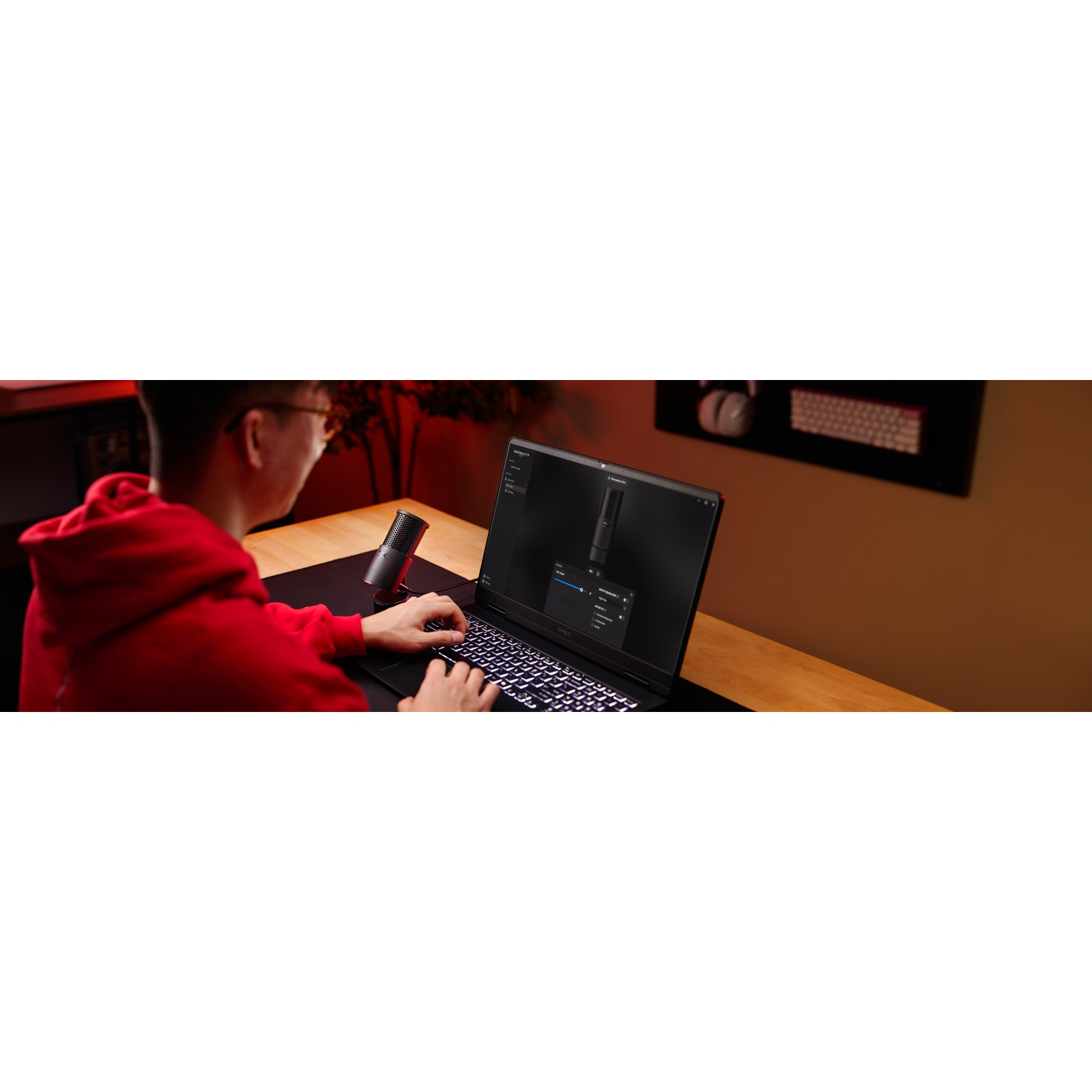 Une personne en sweat à capuche rouge utilise un ordinateur portable avec un microphone à proximité. L'écran affiche des paramètres. Un clavier est visible.