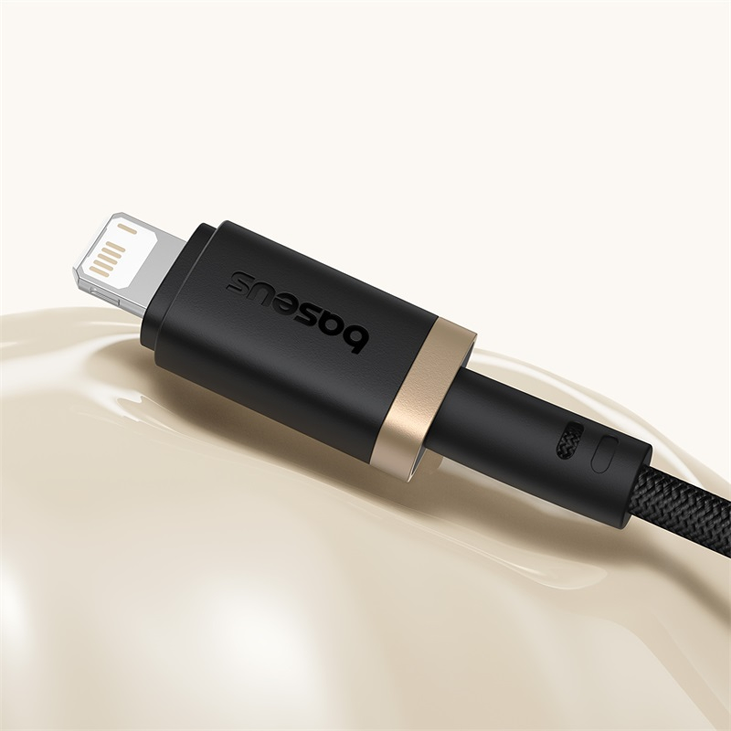 Un cable de carga Baseus negro con un anillo dorado y conector Lightning sobre una superficie beige brillante.