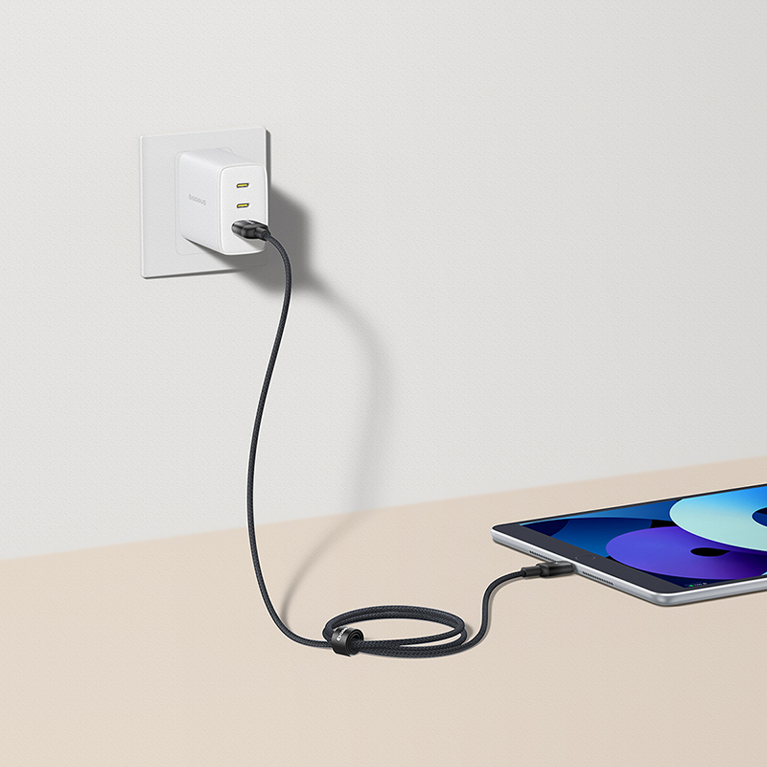 Un cargador blanco está enchufado a una pared, con un cable negro conectado a una tableta sobre una superficie beige.