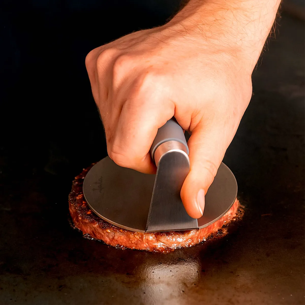 Una mano presiona una hamburguesa con una prensa de metal sobre una superficie caliente. La hamburguesa se está cocinando. El fondo es oscuro.