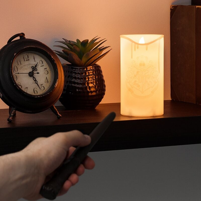 Una mano con una varita señala una vela encendida. Un reloj y una planta están en un estante. La vela tiene un escudo.