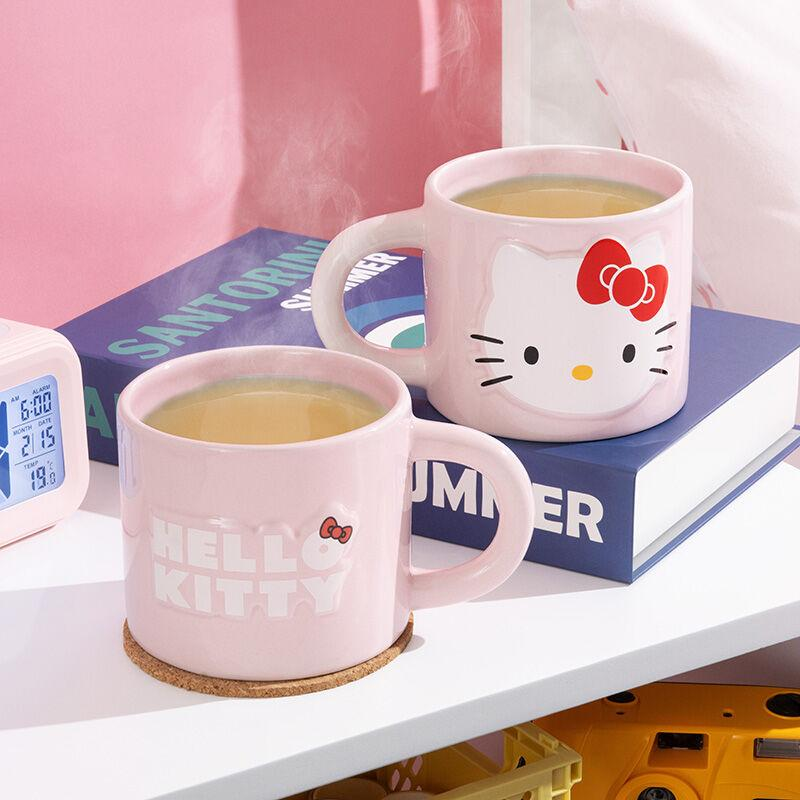 Dos tazas de Hello Kitty con té sobre una repisa blanca. Un libro y un reloj están detrás. Las tazas son rosas con caras de Hello Kitty.