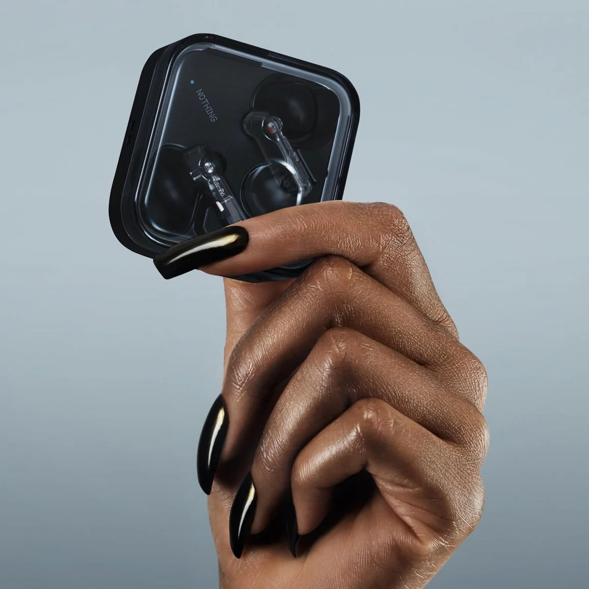 Una mano con esmalte de uñas negro sostiene un estuche transparente con auriculares. El fondo es un degradado de gris.