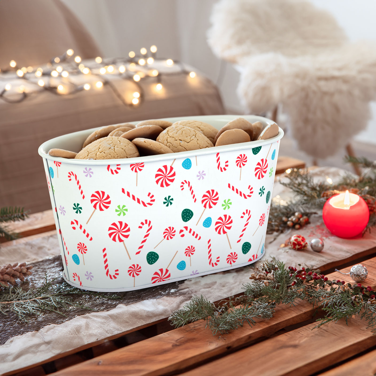 Egy fehér, cukorkákkal díszített, süteményekkel teli doboz egy faasztalon, dekorációkkal, gyertyával és tündérfényekkel a háttérben.