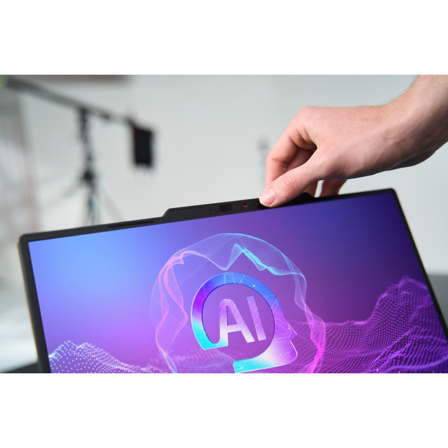 Een hand bedekt de camera van een laptop. Het scherm toont een AI-afbeelding in paarse en blauwe tinten. Er zit een lampje bovenop.