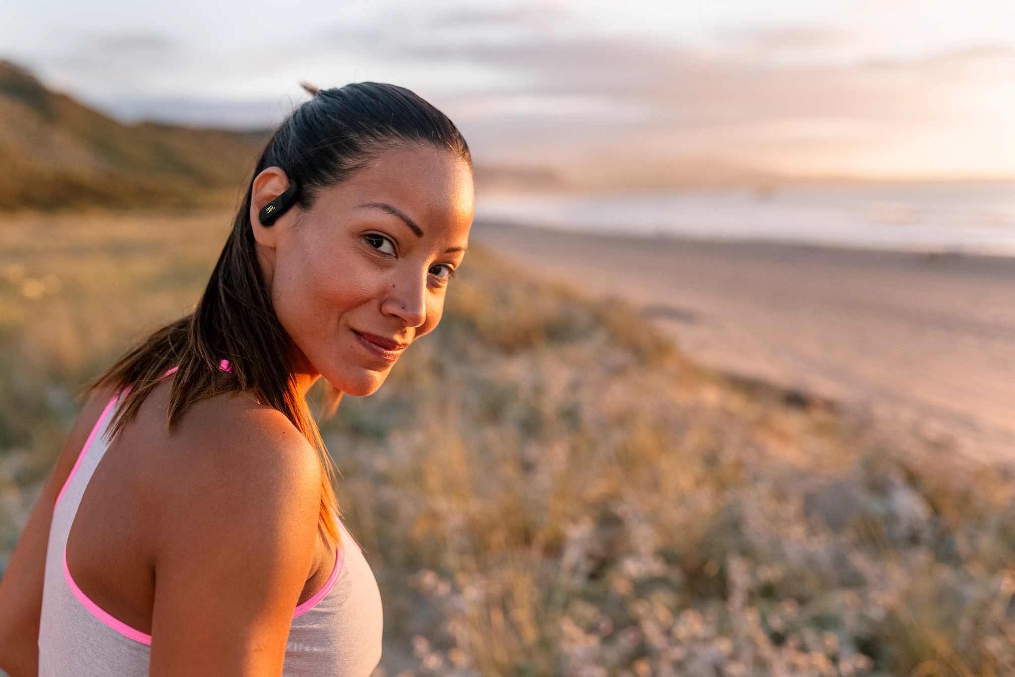 Femme avec écouteurs sourit, regardant la caméra. Plage et dunes en arrière-plan au coucher du soleil.