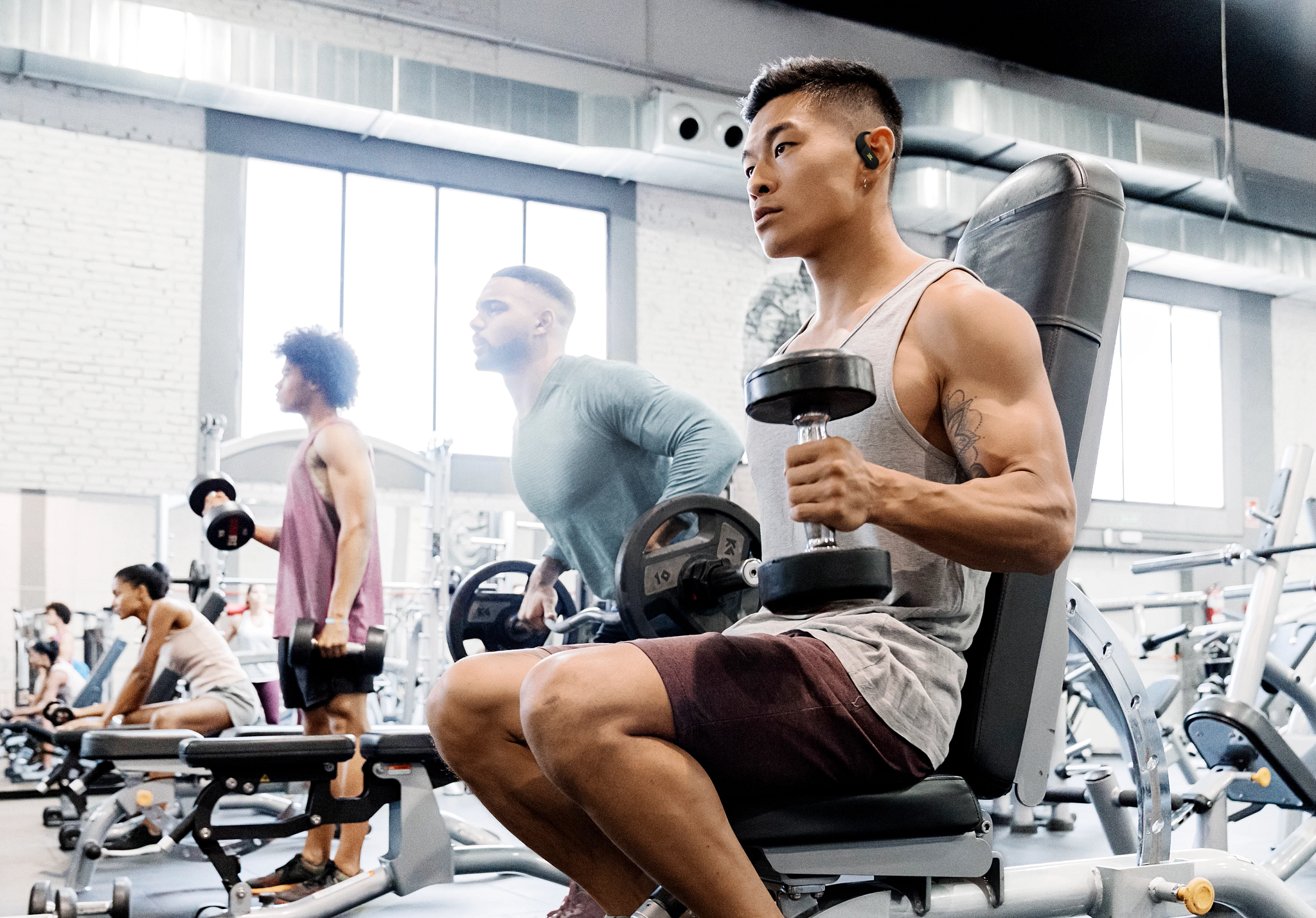 Les gens s'entraînent dans une salle de sport. Un homme soulève un haltère. D'autres s'entraînent avec des poids. La salle de sport a de grandes fenêtres.