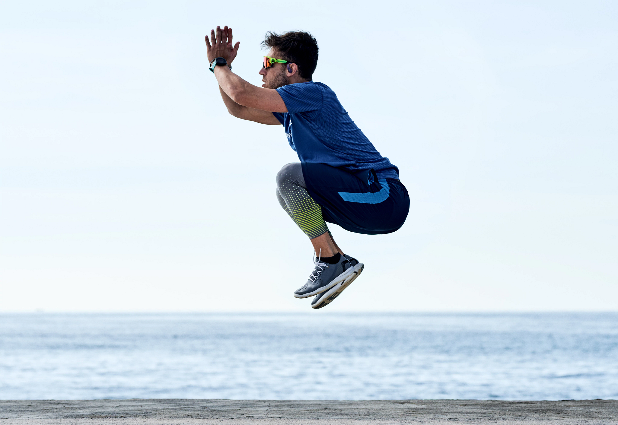 Un homme en tenue de sport saute. Il porte des lunettes de soleil et une montre. La mer et le ciel sont en arrière-plan.