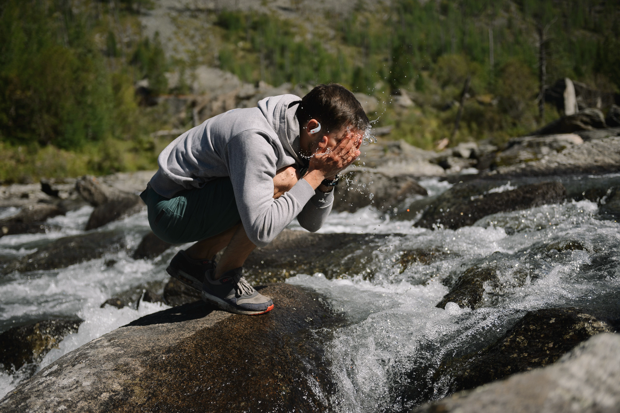 Un homme se lave le visage dans une rivière. Il porte un sweat à capuche gris et un short. L'eau éclabousse alors qu'il se met les mains en coupe.