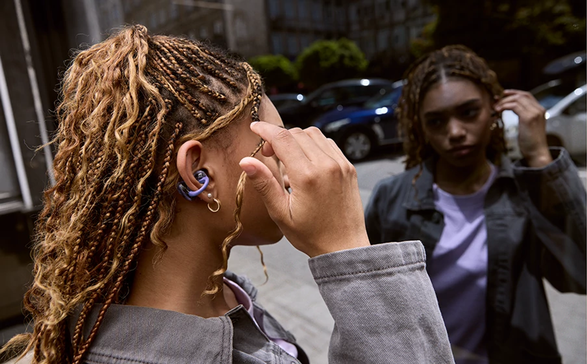 Deux femmes aux cheveux tressés. L'une porte un écouteur, touchant son oreille. L'autre se coiffe. Voitures et bâtiments en arrière-plan.