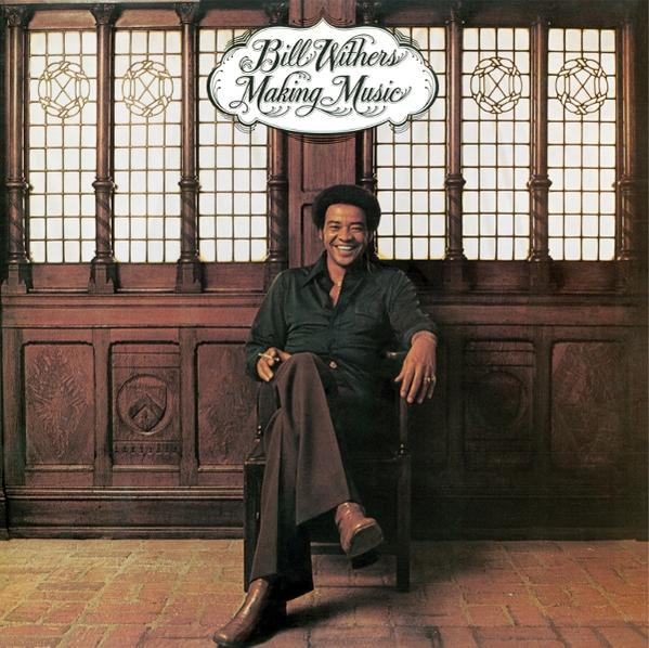 Bill Withers sitzt auf einem Stuhl. Er trägt ein grünes Hemd und braune Hose. Der Hintergrund ist eine Holzwand und Fenster.