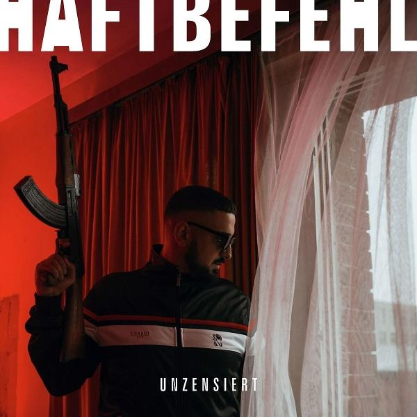 Mann mit Waffe, Sonnenbrille und schwarzer Jacke. Rote Vorhänge und Fenster mit weißen Vorhängen im Hintergrund.