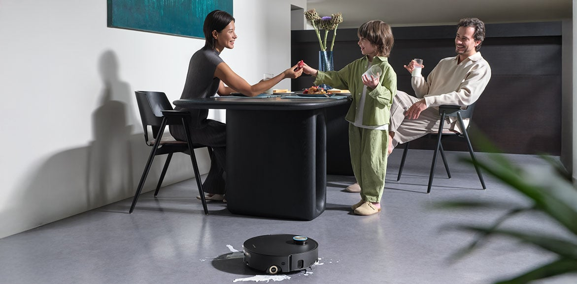 Rodzina przy stole, dziecko częstuje jedzeniem. Robot sprzątający na podłodze. Nowoczesne wnętrze z obrazem i roślinami.