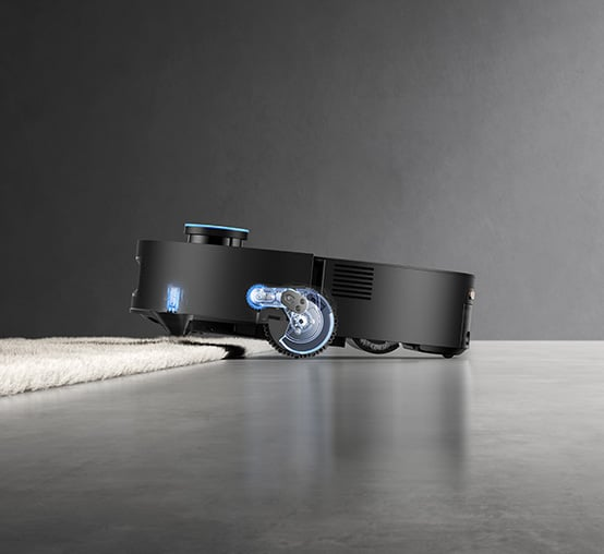 Czarny robot sprzątający na szarej podłodze, przechodzący w biały dywan. Odkurzacz ma niebieskie światło.