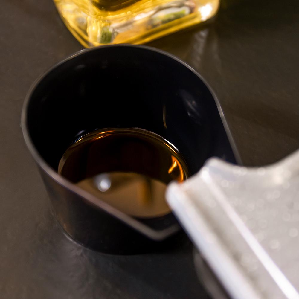 Una taza medidora negra contiene líquido oscuro, posiblemente aceite. Una botella de vidrio de aceite está borrosa en el fondo, sobre una superficie oscura.